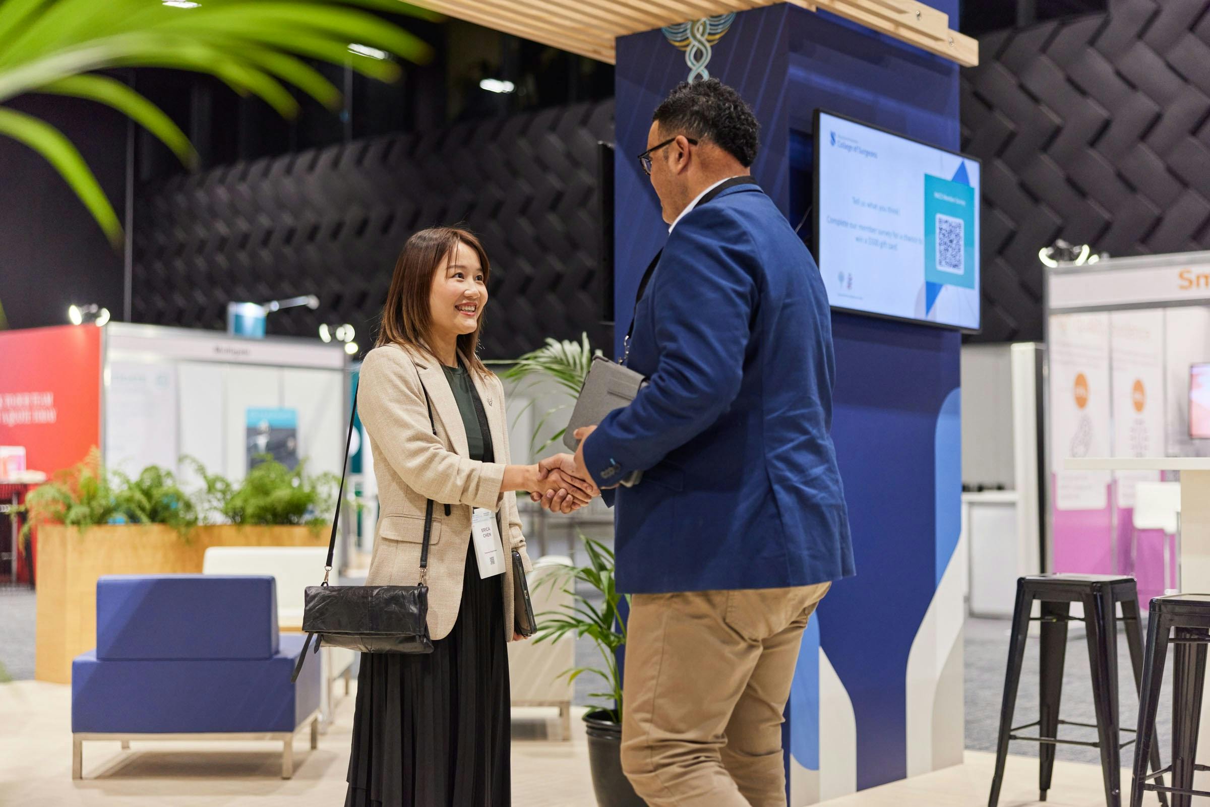 Two professionals are shaking hands in a modern exhibition space featuring Te Pae branding, surrounded by greenery and booths with informational displays in the background.